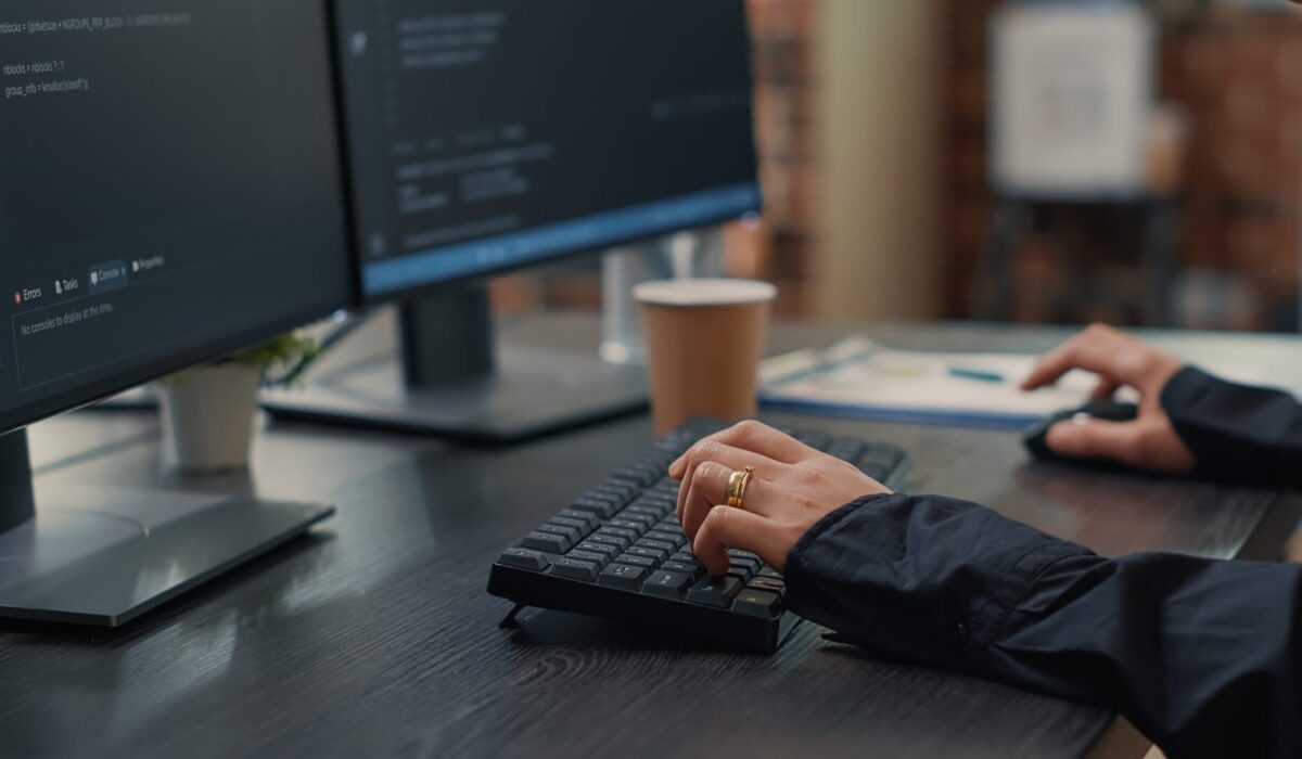 Closeup of developer hands typing code on keyboard while looking at computer screens with programming interface. Software programmer sitting at desk with clipboard writing algorithm.